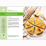 Eine Rezeptseite aus dem Fettverlust Fibel Backbuch zeigt einen in Scheiben geschnittenen Eiweiß-Kuchen mit Apfelspalten auf einem Teller, umgeben von Weizen, einem halben Apfel, einem gestreiften Tuch und einem deutschen Rezept mit Zutaten und Anleitung.