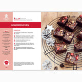 Eine Rezeptseite aus dem Fettverlust Fibel Backbuch zeigt schokoladenüberzogene Kuchenquadrate mit goldenen Streuseln und roten Beeren auf einem Drahtgestell, mit Tannenzapfen und sternförmigen Ornamenten in der Nähe für eine verlockende, gesunde Desserts-Auslage.