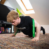Ein Mann mit lockigem Haar macht Liegestütze mit dem Widerstands-Bänder Set (+ Gratis Buch) auf einem Teppich. Er trainiert zu Hause in einem hellen Raum mit einem Fernseher, Büchern und einer Pflanze - perfekt für sein persönliches Trainingsprogramm.
