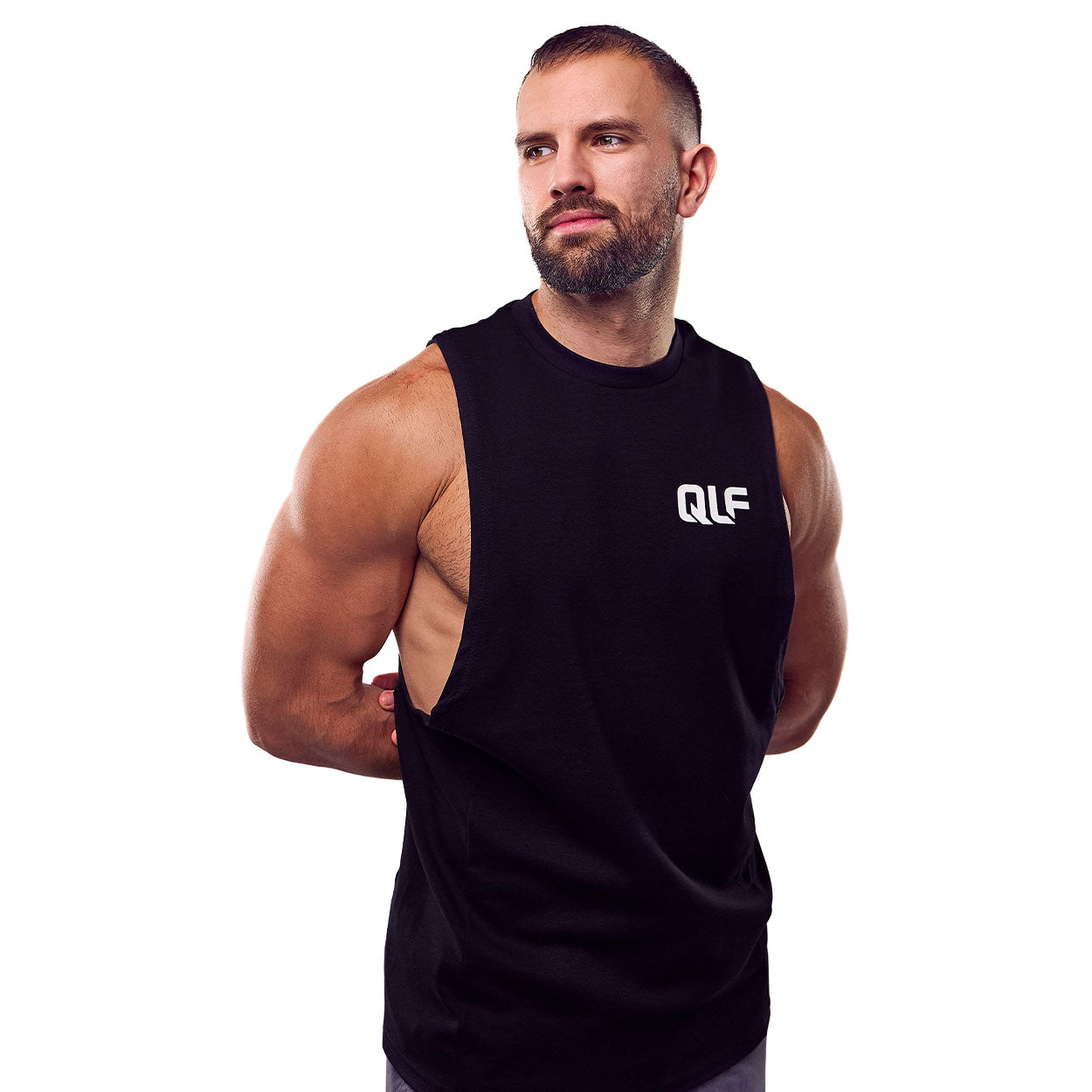 Ein bärtiger, muskulöser Mann trägt ein ärmelloses schwarzes QLF Squad Tank Top (kleines Logo). Er steht vor einem weißen Hintergrund und hat die Arme hinter dem Rücken verschränkt, wobei er seine Muskeln betont und leicht zur Seite blickt.