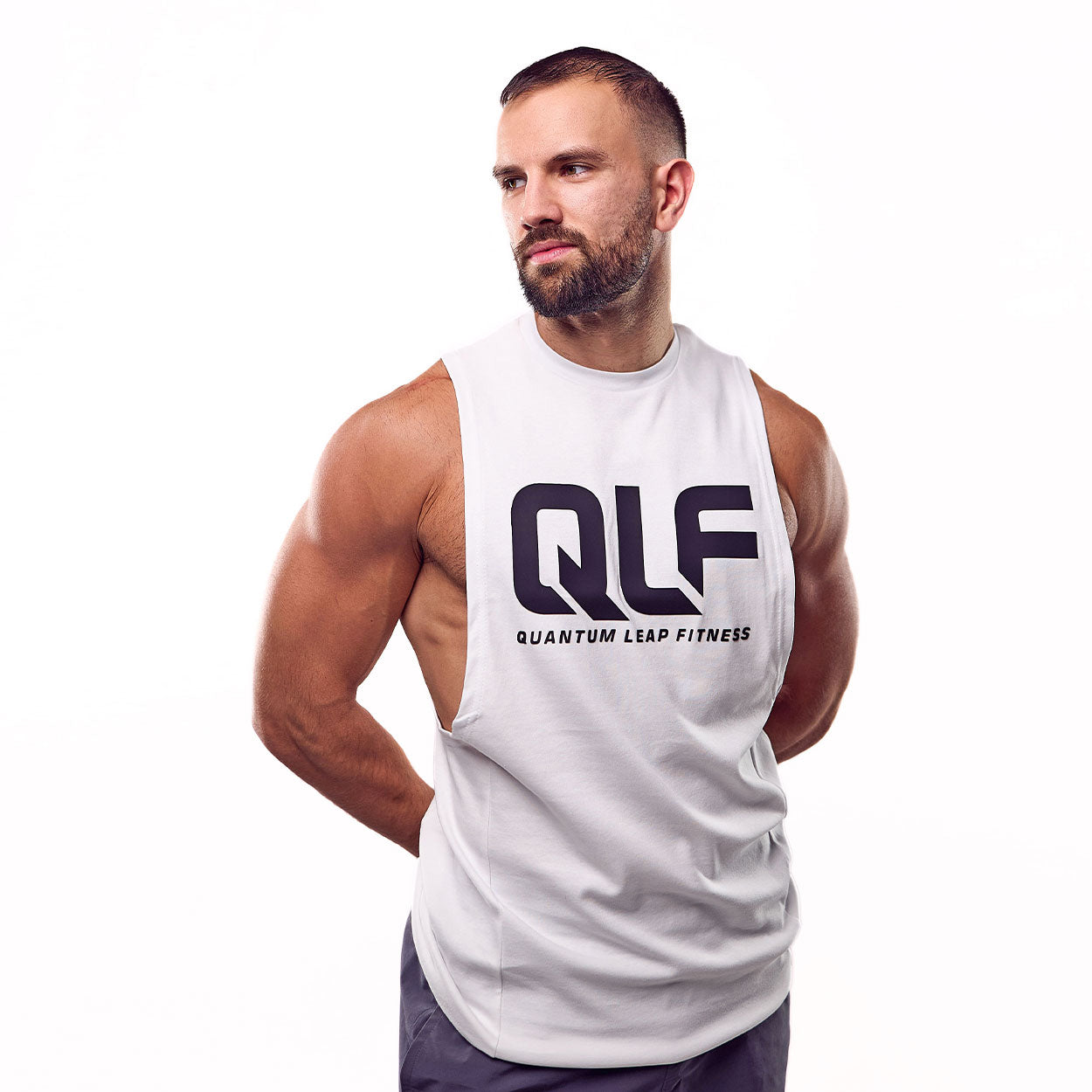 Ein muskulöser Mann mit Bart steht vor einem weißen Hintergrund und trägt das QLF Squad Tank Top (großes Logo). Er blickt mit den Händen hinter dem Rücken zur Seite und zeigt das ideale Gym Muskelshirt für Fitnessbegeisterte.