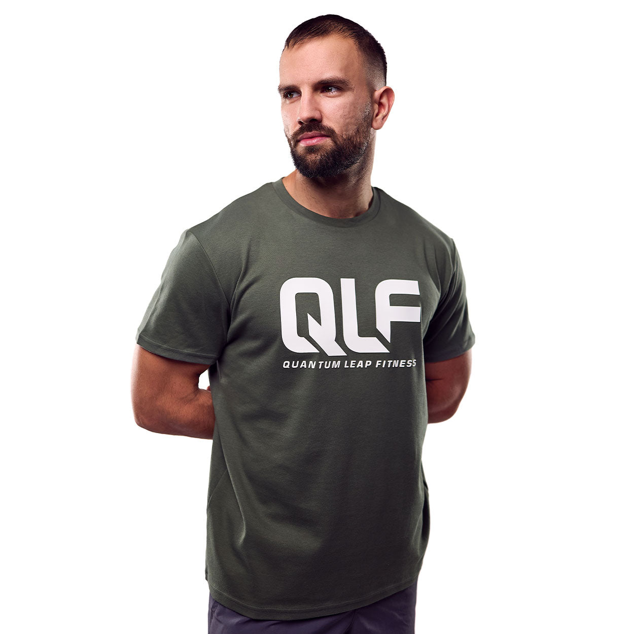 Ein bärtiger Mann steht selbstbewusst mit den Händen auf dem Rücken und trägt das olivgrüne QLF Squad T-Shirt (großes Logo) mit einem auffälligen weißen QLF Logo und Quantum Leap Fitness Text auf der Vorderseite.