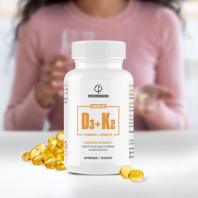 Eine Flasche mit D3+K2-Vitaminpräparaten wird auf einem Tisch gezeigt, auf dem gelbe Kapseln verstreut sind. Im unscharfen Hintergrund hält eine Person ein Glas Wasser und eine Kapsel.