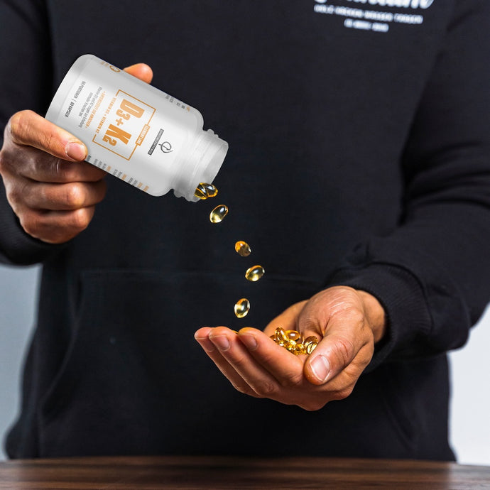 Eine Person in einem schwarzen Kapuzenpulli schüttet gelbe Softgel-Kapseln aus einer weißen Flasche mit der Aufschrift "D3 + K2" in ihre Hand auf eine Holzfläche.