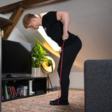 Eine Person in schwarzer Trainingskleidung trainiert mit dem Widerstands-Bänder Set (+ Gratis Buch) in einem gemütlichen Wohnzimmer mit Fernseher, Pflanzen, Büchern und schräger Decke - ideal für ein persönliches Trainingsprogramm zu Hause.