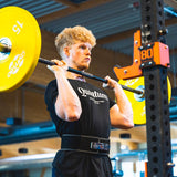 Ein Mann mit lockigem blondem Haar, der ein schwarzes Quantum Leap Fitness-T-Shirt und einen Gewichthebergürtel trägt, führt im Fitnessstudio eine Kniebeuge mit einer gelben 15-kg-Hantel durch.