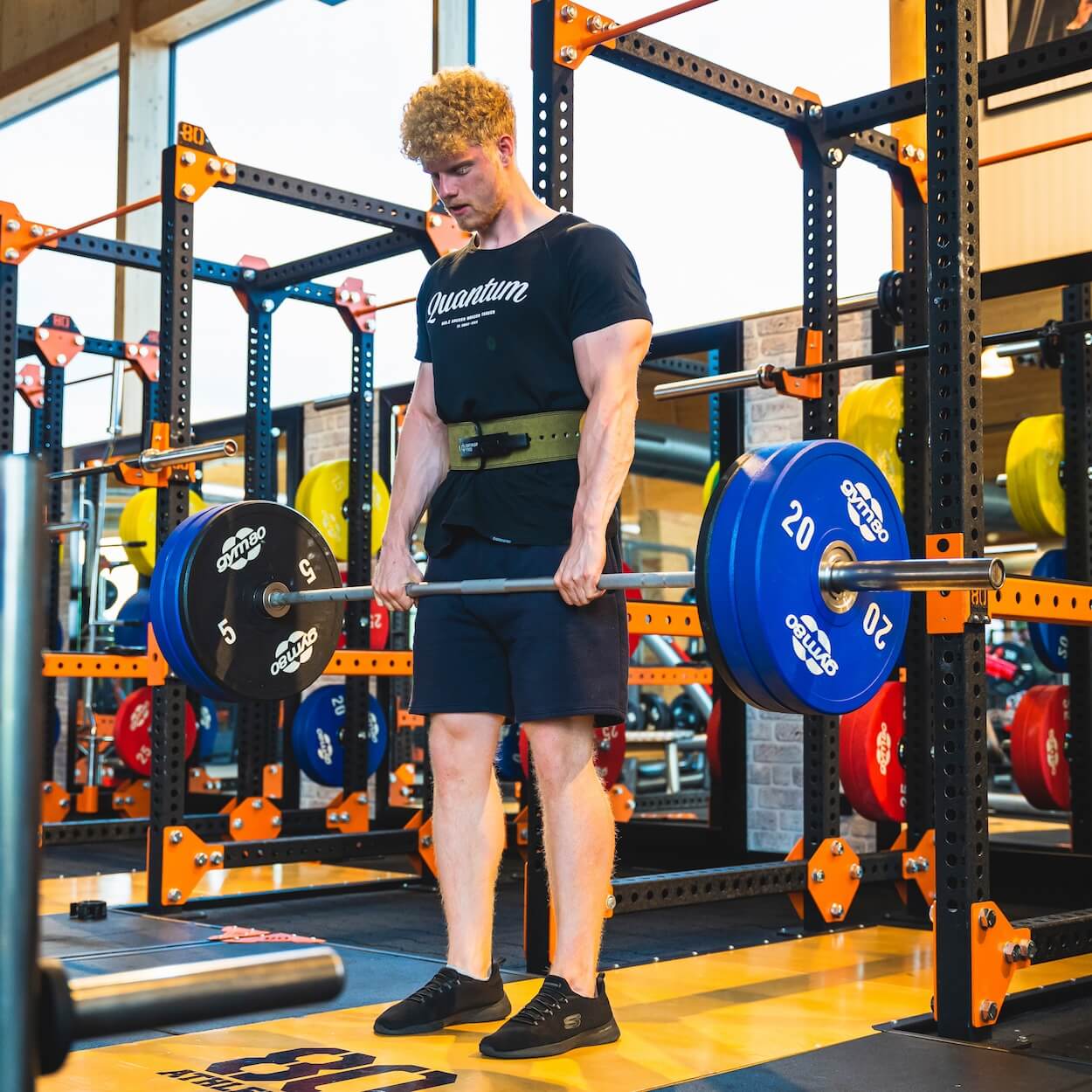 Ein Mann in schwarzem T-Shirt und Shorts hebt im Fitnessstudio Quantum Leap Fitness eine belastete Langhantel mit einem Gewichthebergürtel. Er steht auf einer Plattform, konzentriert auf Grundübungen, mit Fitnessgeräten und Gewichten im Hintergrund.