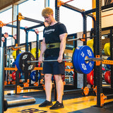 Ein Mann in schwarzem T-Shirt und Shorts hebt im Fitnessstudio Quantum Leap Fitness eine belastete Langhantel mit einem Gewichthebergürtel. Er steht auf einer Plattform, konzentriert auf Grundübungen, mit Fitnessgeräten und Gewichten im Hintergrund.