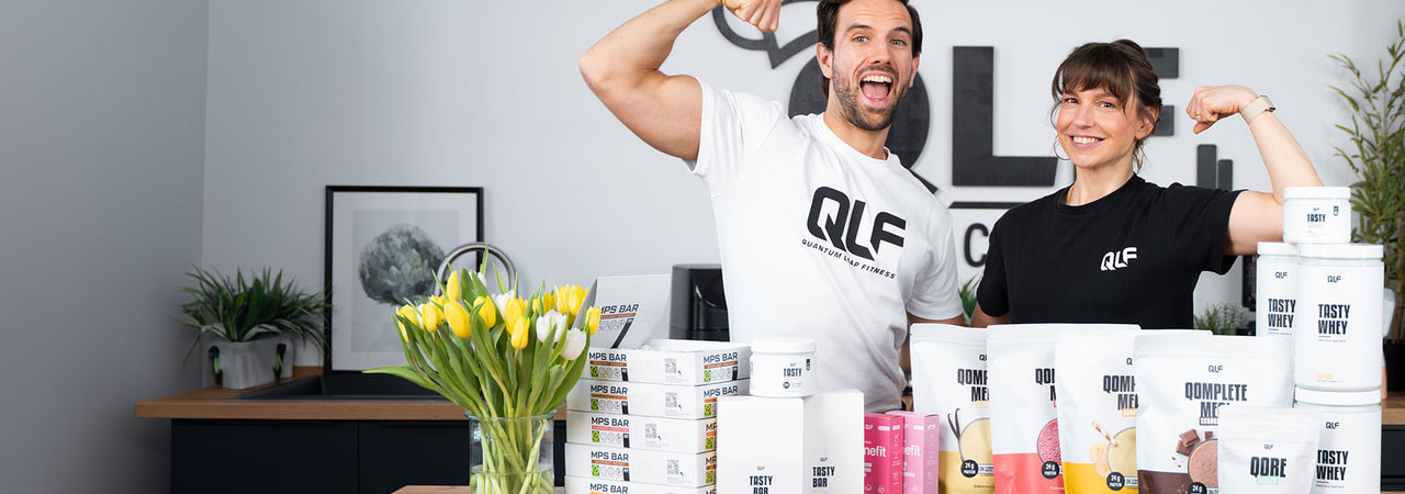 Zwei Personen mit QLF-Shirts zeigen lachend Muskelkraft hinter einem Tisch mit Proteinpulver und Stoffwechselshakes