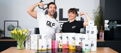 Zwei Personen mit QLF-Shirts zeigen lachend Muskelkraft hinter einem Tisch mit Proteinpulver und Stoffwechselshakes
