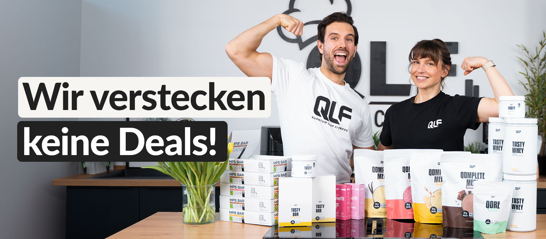 Zwei Personen mit QLF-Shirts zeigen zufrieden und selbstbewusst Muskelmuskeln neben gestapelten Verpackungen und Bechern