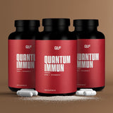 Drei Quantum-Immun-Dosen mit roten Etiketten, die Vitamin C und Zink zur Unterstützung des Immunsystems enthalten, sind nebeneinander angeordnet, mit weißen Kapseln und Pulver davor, vor einem braunen Hintergrund.