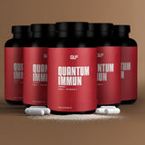 Sechs Quantum-Immun-Dosen mit roten Etiketten sind in zwei Reihen angeordnet. Vorne sind weiße Kapseln und Tabletten mit hohem Zink- und Vitamin-C-Gehalt zur Unterstützung des Immunsystems auf einer braunen Fläche vor einem einfarbig braunen Hintergrund ausgestellt.