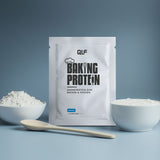 Ein Backproteinpaket liegt zwischen einer Schale mit weißem Pulver, einer Schale mit Joghurt und einem Löffel - bereit für Fitnessrezepte mit perfekter Konsistenz - alles vor einem blauen Hintergrund.