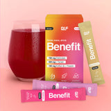 Ein Glas mit rotem Getränk steht neben einer farbenfrohen Benefit-Energy-Box mit Angaben zu Nahrungsergänzungsmitteln und Hydration-Formel sowie drei Portionsbeuteln in Pink, Lila und Gelb, die auf einer rosafarbenen Fläche vor einem Hintergrund mit Farbverlauf präsentiert werden.