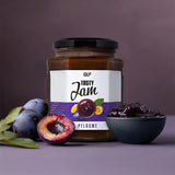 Ein Glas Tasty Jam Pflaume wird zusammen mit frischen Pflaumen, einer aufgeschnittenen Pflaume und einer Schale mit dunkler Pflaumenmarmelade - einem unwiderstehlichen kalorienarmen Fruchtaufstrich mit 80 % Fruchtanteil - auf einem leuchtend violetten Hintergrund gezeigt.