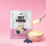 Eine Packung High Protein Tasty Pudding steht vor einem rosafarbenen Hintergrund neben einer Schüssel mit Pudding. Ein Löffel hebt die proteinreiche Leckerei an, in der Nähe liegen frische Blaubeeren, die den Muskelaufbau unterstützen.