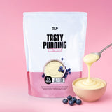 Eine Packung High Protein Tasty Pudding mit einer Schale Pudding, Blaubeeren und einem Löffel, der den Pudding ausgießt. Der rosa Hintergrund enthält 19 g Protein, 87 kcal und 1,9 g Kohlenhydrate pro Portion - ideal für den Muskelaufbau.