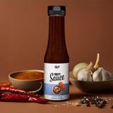 Eine Flasche Tasty Sauce steht zwischen einer Schale mit Soße, roten Chilischoten, Knoblauchknollen und verstreuten Pfefferkörnern auf einer braunen Fläche mit braunem Hintergrund, was ihre Attraktivität als kalorienarme Diätzutat unterstreicht.