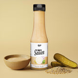 Eine Flasche Tasty Sauce steht neben einer Schale mit der kalorienarmen Sauce, in Scheiben geschnittenen Essiggurken und Senfkörnern, die alle auf einem beigen Hintergrund angeordnet sind.