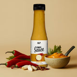 Eine Flasche Tasty Sauce ist neben roten Chilischoten, Knoblauchzehen, Petersilie und einer kleinen Schale mit der kalorienarmen Sauce mit einem Löffel darin auf einem beigen Hintergrund zu sehen.
