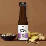 Eine Flasche Tasty Sauce steht auf einer braunen Fläche neben einer Schale mit gemischten Sesamsamen und frischen Ingwerscheiben. Diese kalorienarme Sauce ist ideal für gesunde Mahlzeiten und steht vor einem soliden braunen Hintergrund.