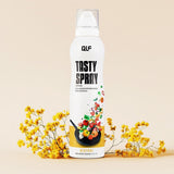 Eine Dose Tasty Spray steht aufrecht auf einer beigen Fläche inmitten kleiner gelber Blumen. Auf dem Etikett sind Gemüse und Kräuter abgebildet, die in eine Bratpfanne fliegen, um die kalorienarme Formel und die genaue Dosierung zu unterstreichen.