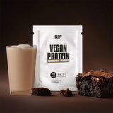 Ein Glas cremiger Vegan-Protein-Shake steht neben einer weißen Vegan-Protein-Packung, mit Schokoladen-Brownies und Krümeln auf einer dunklen Oberfläche - perfekt für Ihre vegane Revolution.
