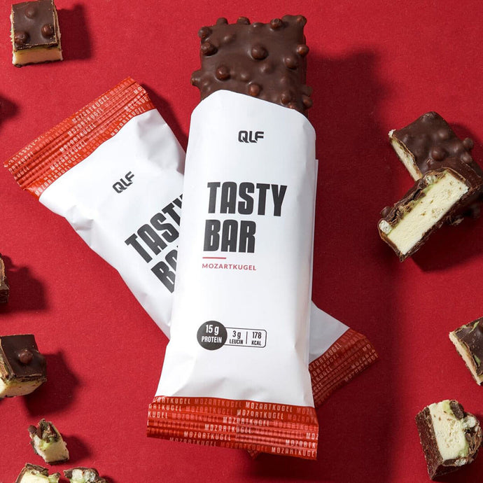 Zwei Tasty Bar Proteinriegel in weißer und roter Verpackung mit Schokoglasur und Nussstücken. Angeschnittene Riegel zeigen die cremige Füllung - der ideale kalorienarme Snack für alle Fitness-Fans.
